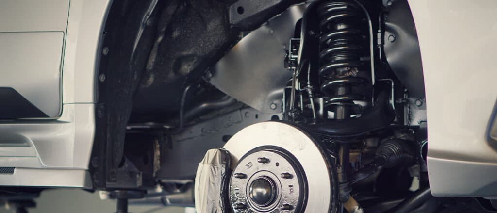 Suspension Repair Signs - C&M Auto Service Expert Advice | Morgan Hill. Image of white car on lift in shop with tire removed so you can see the brakes and suspension.