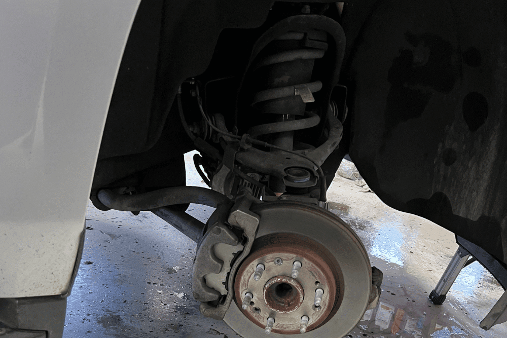 Suspension checkup, auto repair in Morgan Hill, CA by C&M Auto Service, Inc. A close-up view of a car’s front wheel well showing the brake rotor, caliper, suspension coil spring, and control arm with the wheel removed. The vehicle is in a workshop setting.