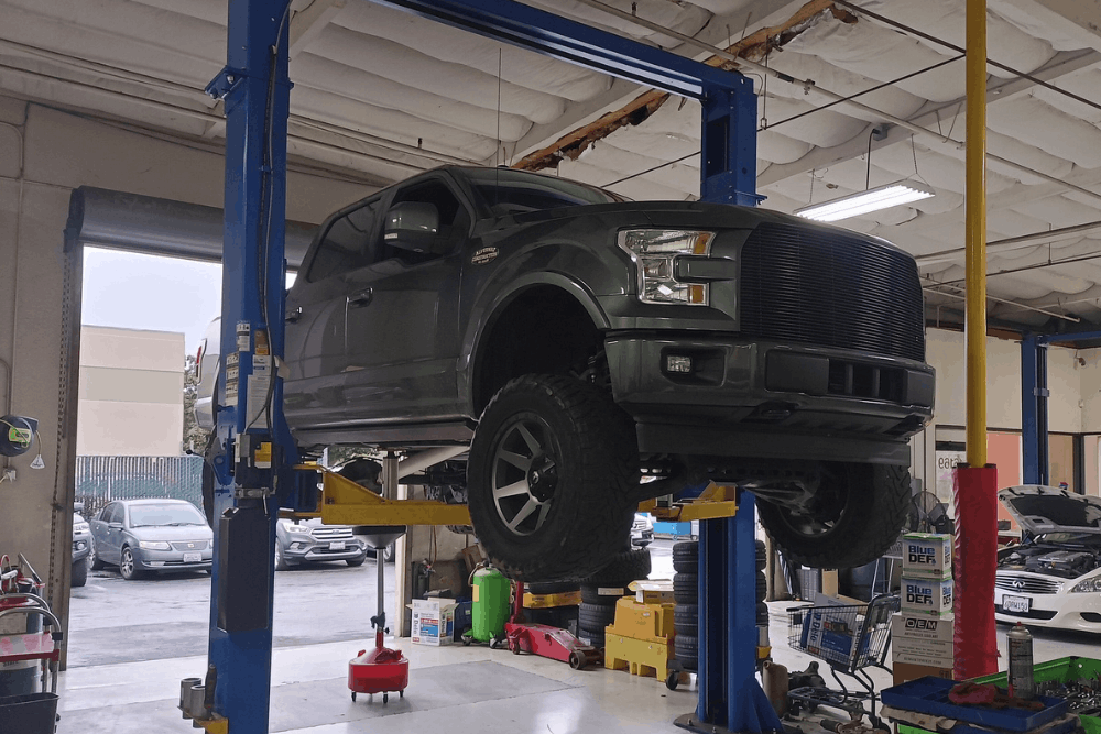 A dark green pickup truck is elevated on a hydraulic lift inside an auto repair shop, surrounded by tools and equipment.
